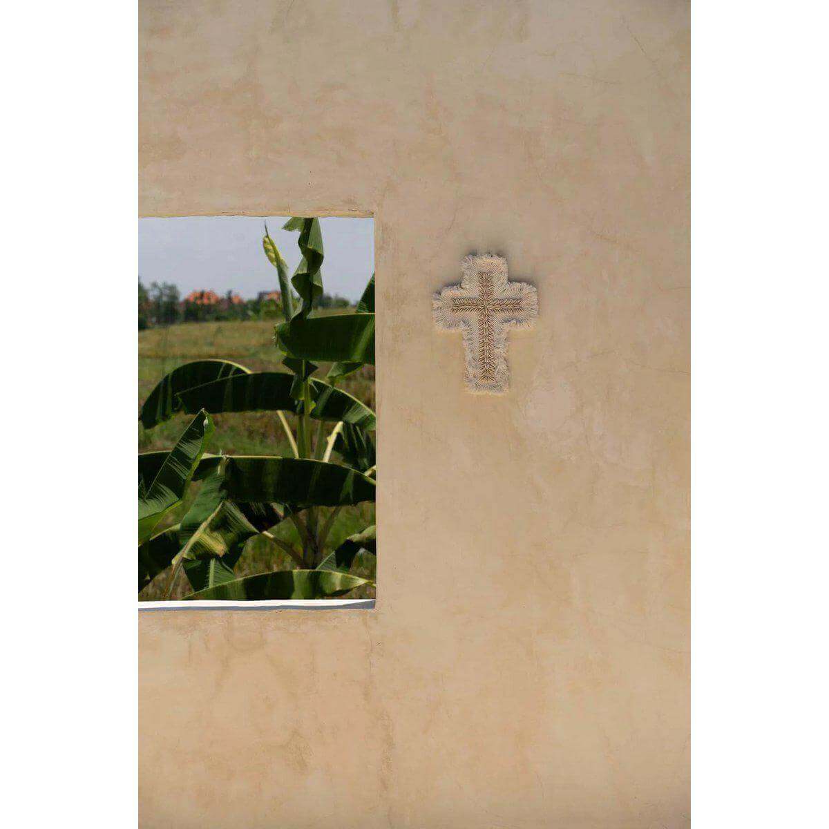 Handgemaakt wit macrame-kruis met kaurischelpen, katoenen franjes en een strandvibe, hangend aan een muur met uitzicht op de natuur.