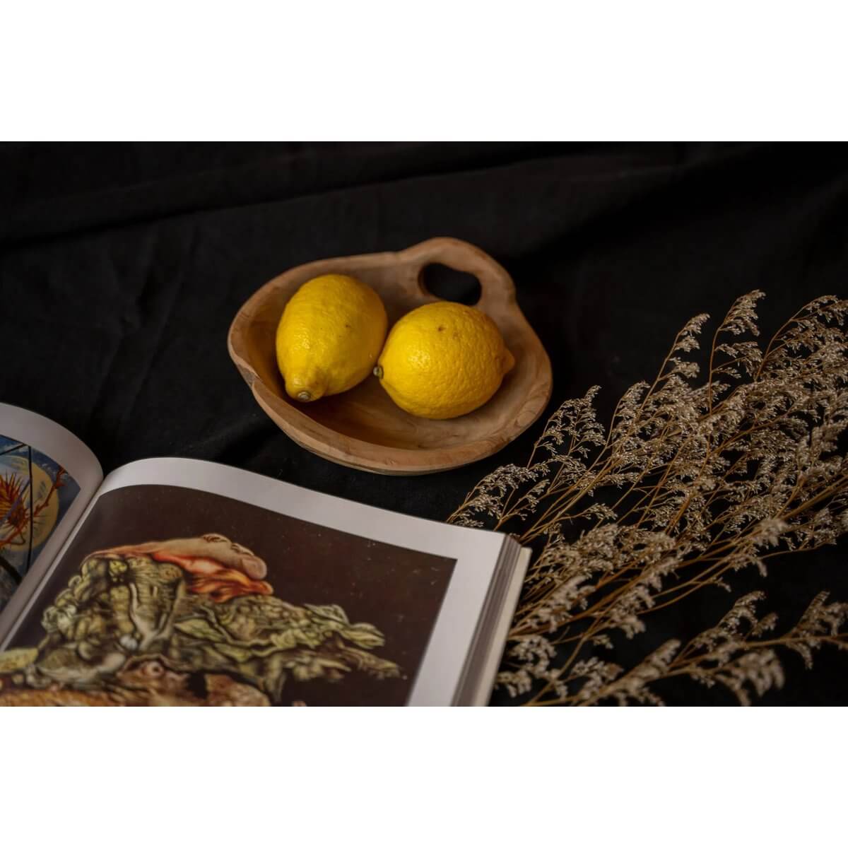 Twee citroenen op houten schaal, naast een open boek en droogbloemen, alle op een zwarte achtergrond.
