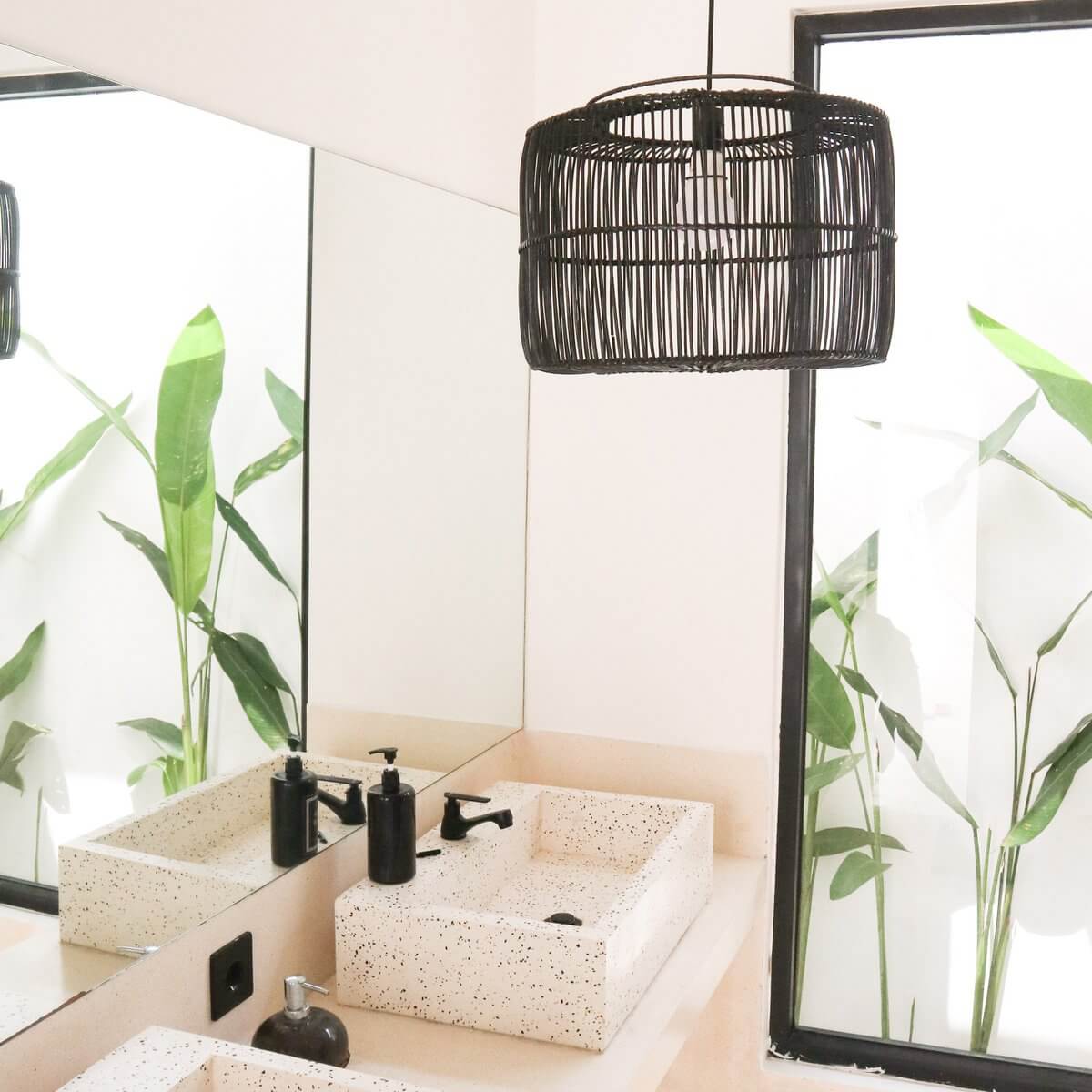 Zwarte rotan plafondlamp handgemaakt, creëert warme patronen, hangt in moderne badkamer met natuurlijk licht en groen blad.