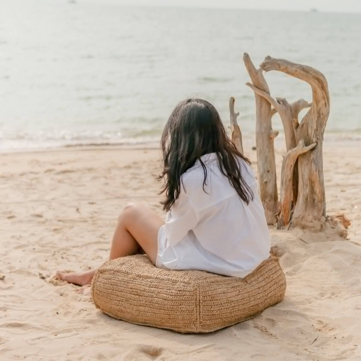 Vrouw zittend op een handgemaakte raffia poef aan het strand, genietend van de rustige zee en zon.
