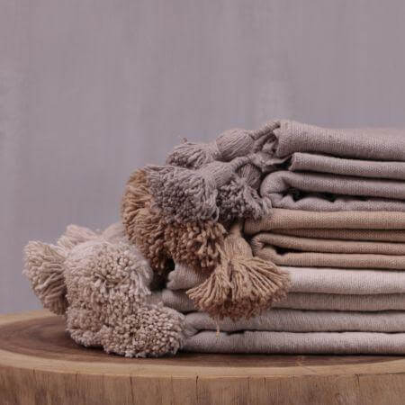 Stapel van beige pompom dekens op houten tafel, perfect voor een modern of boho interieur en gezellige avonden thuis.
