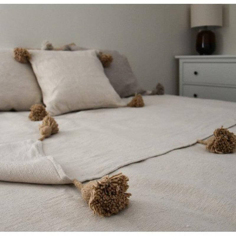 Marokkaanse pompomdeken in crème en beige op een bed, perfecte aanvulling voor elke woonstijl, van modern tot bohemien.