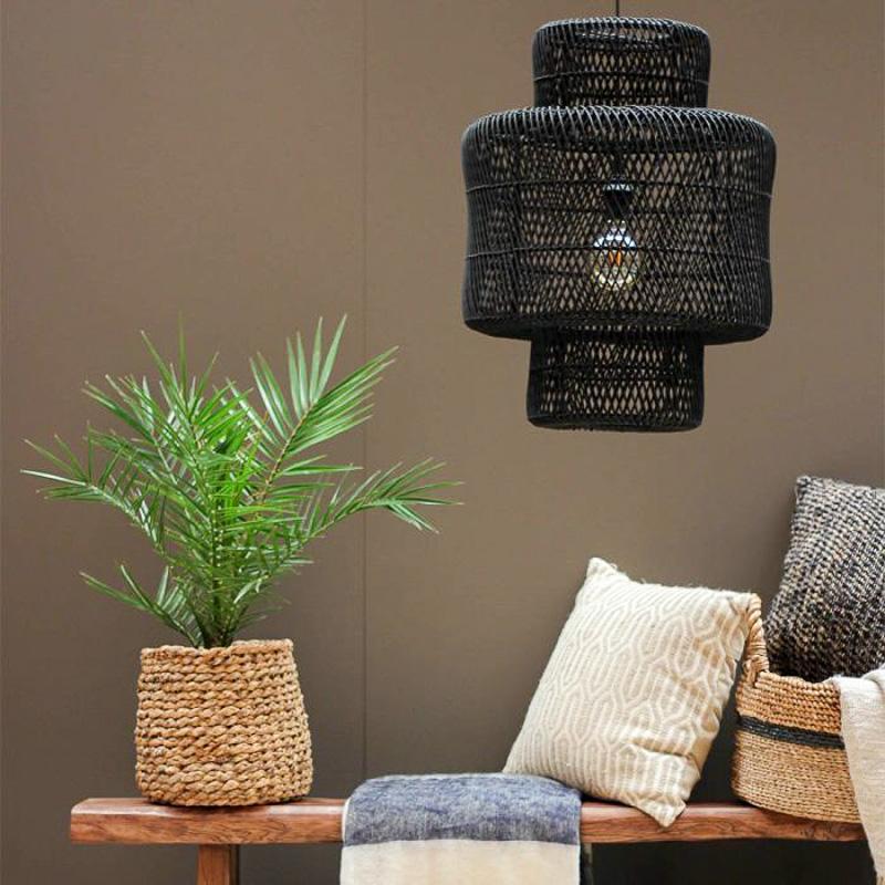 Zwarte rotan hanglamp Janine hangt boven een houten tafel met groen plantje en kussens, sfeervol interieurontwerp idee.
