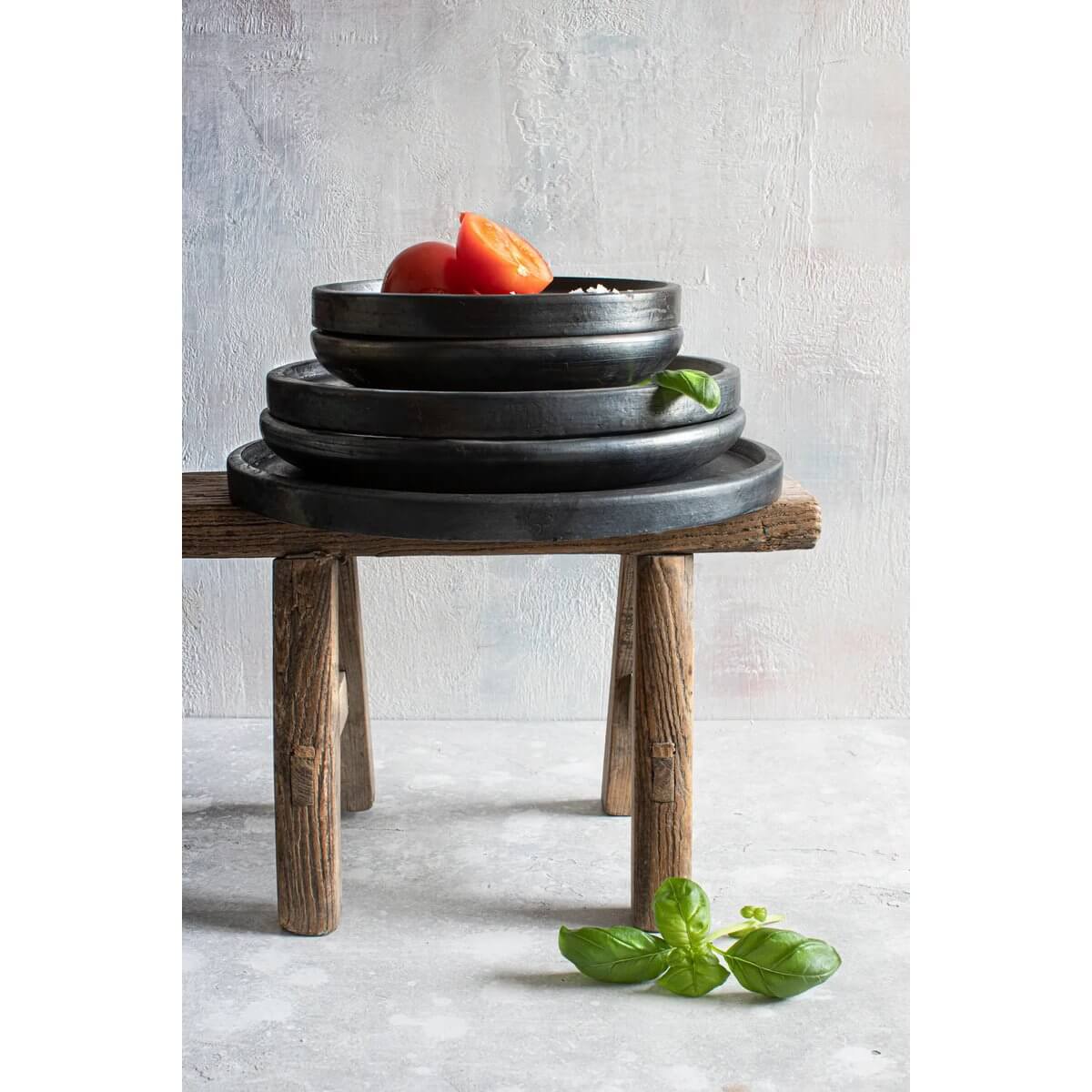Stacked black terracotta plates on wooden stand with basil and tomatoes, showcasing Bohemian style. Width 20cm, by Bazar Bizar Living.
