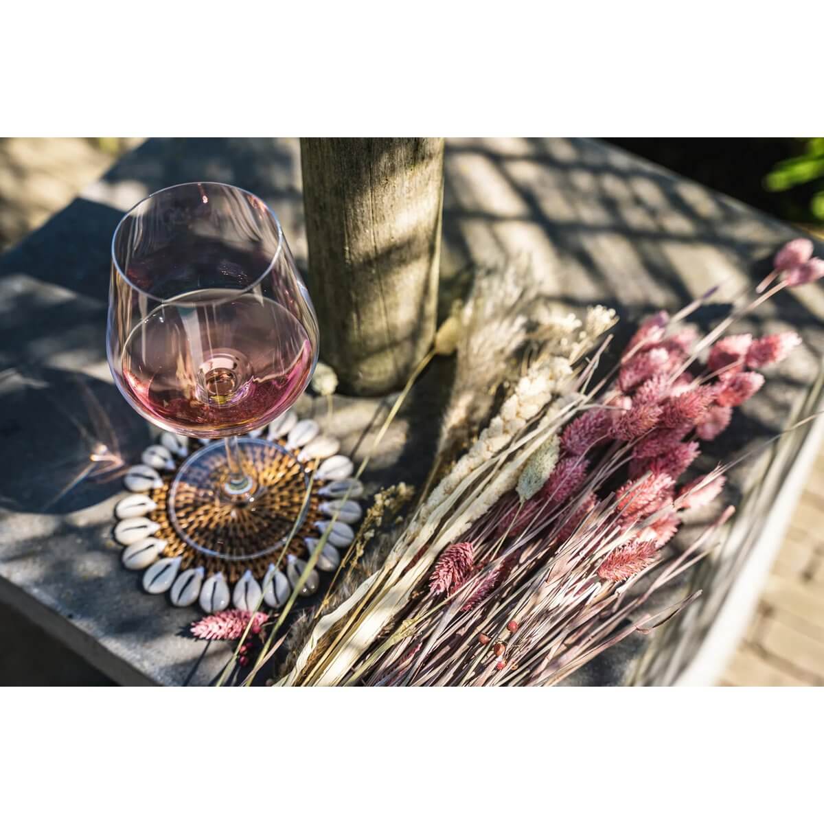 Bohemian style glass of wine on a Colonial Shell Coaster with rattan and kauri shells, next to dried flowers, adding a tropical island touch.