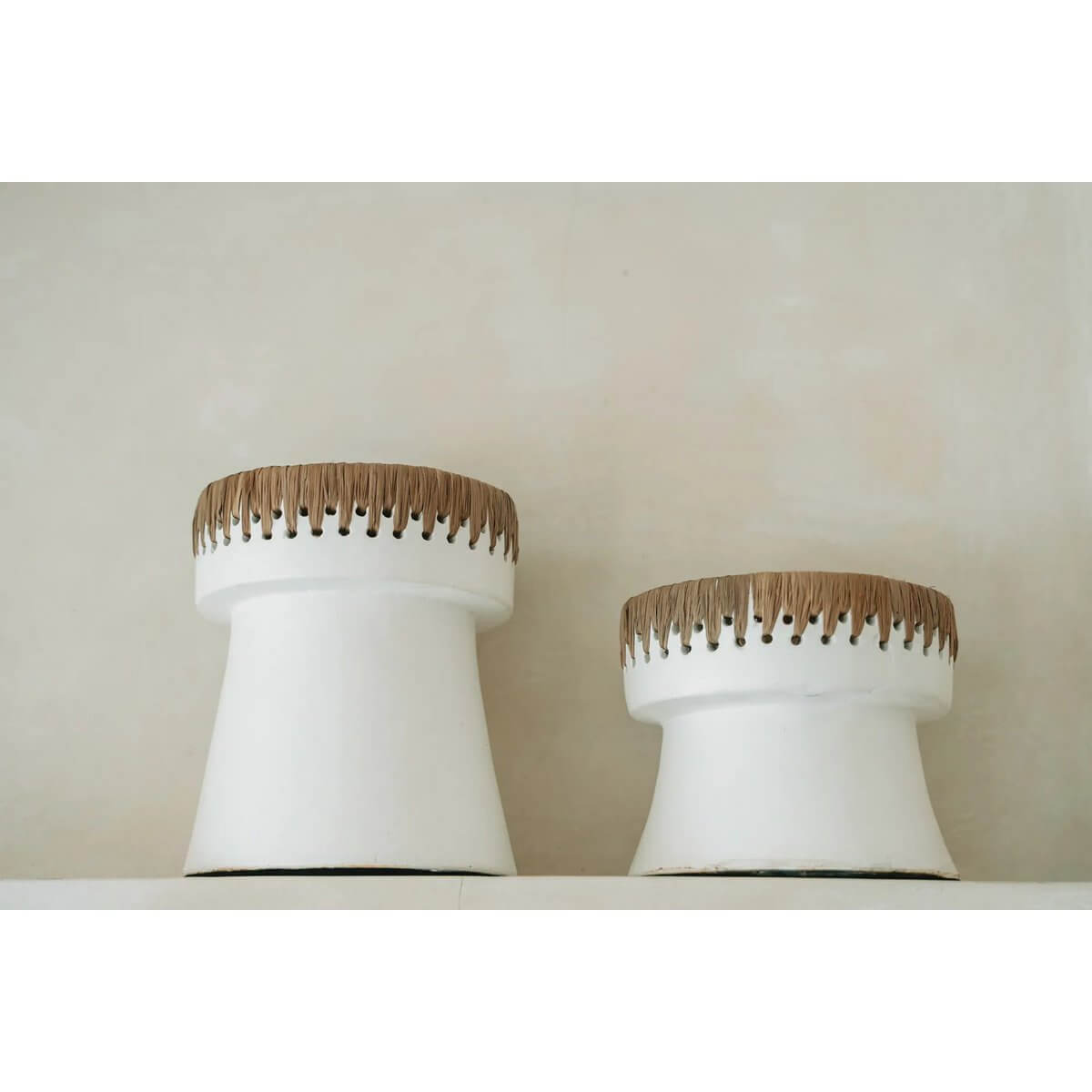 Two white terracotta and raffia bohemian candle holders in varying heights on a shelf, perfect for adding style to any space.