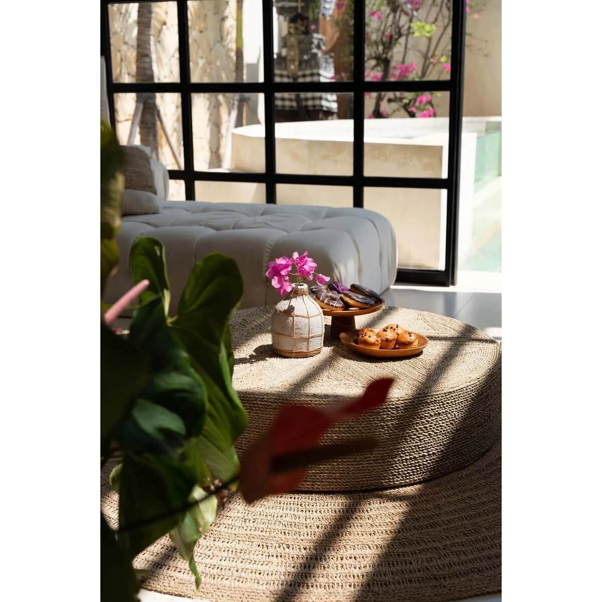 Gezellige woonkamer met teakhouten gebaksborden en bloemen op een rotan salontafel in zonlicht.