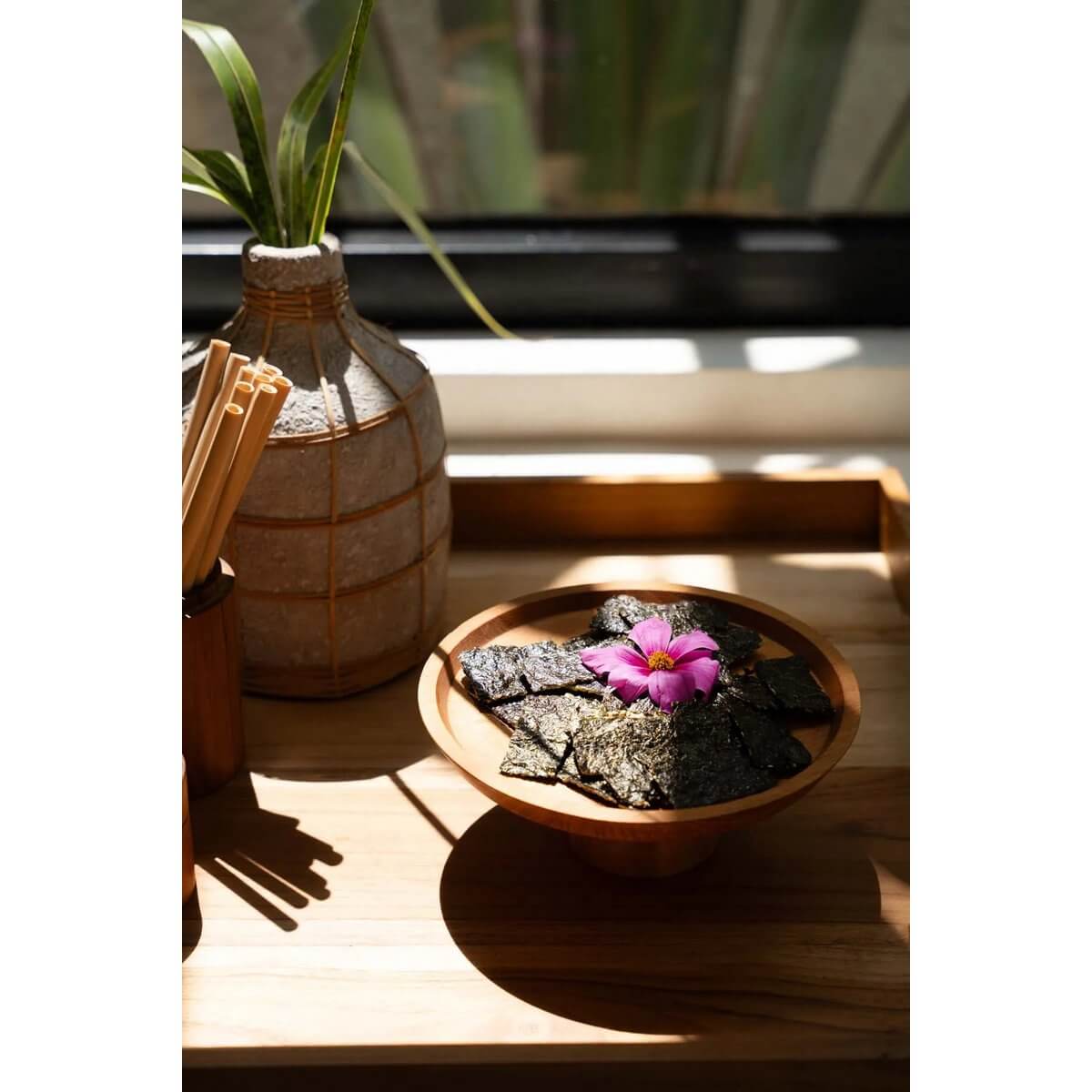 Bohemian teak root cake dish with snacks and a pink flower, styled on a wooden tray beside a decorative vase and chopsticks.