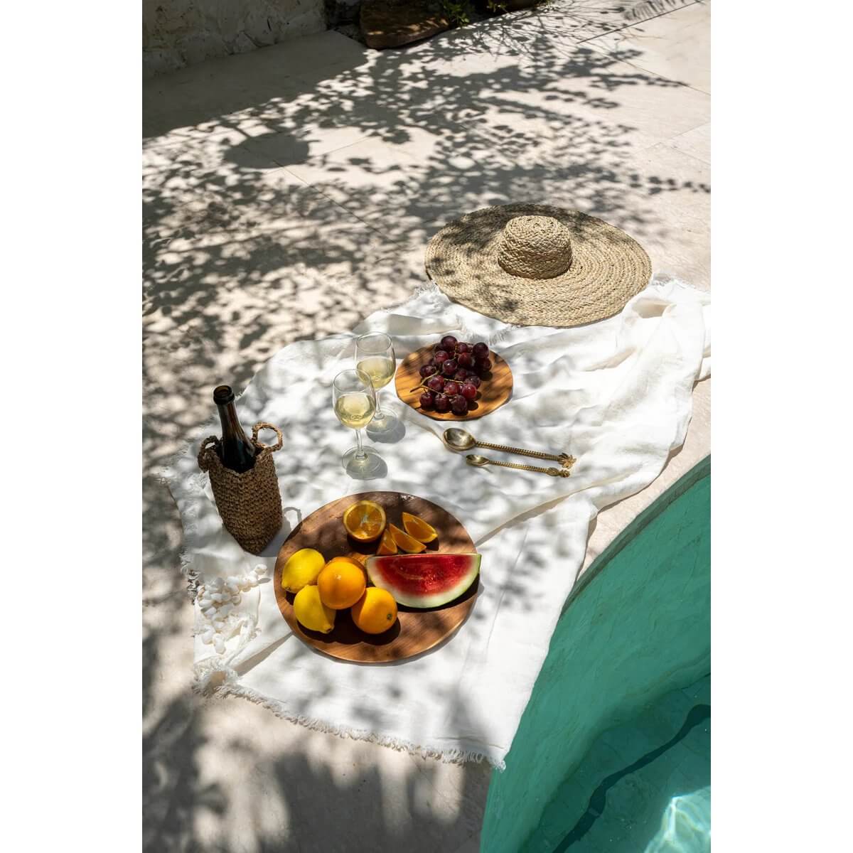 Picknick bij het zwembad met fruitschaal, wijn en hoed op wit kleed.