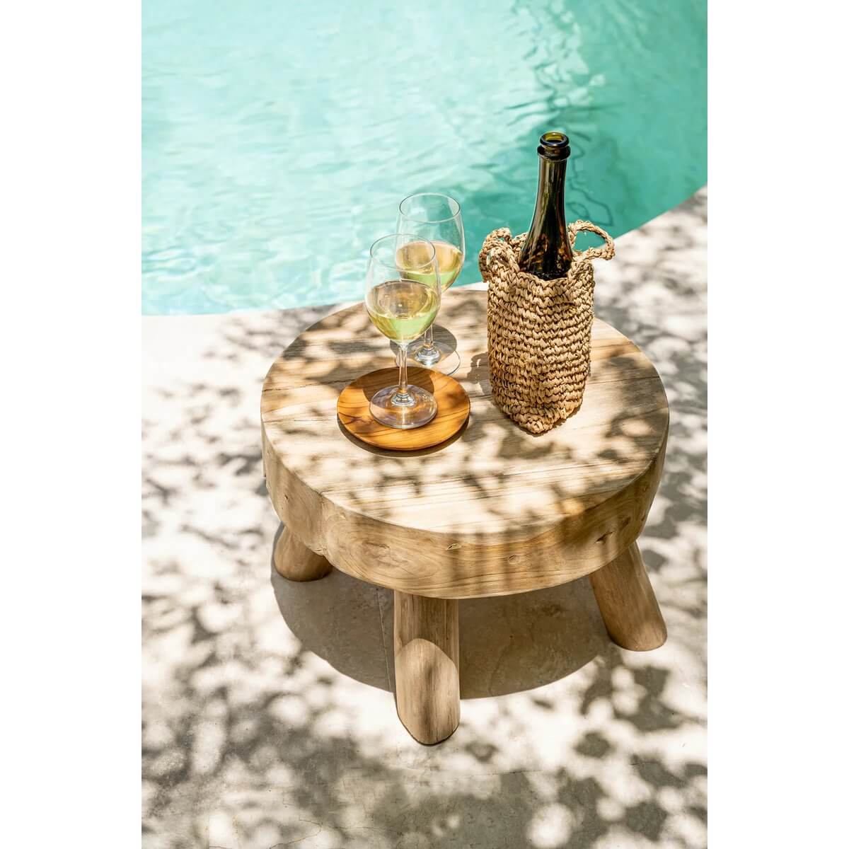 Sunny poolside scene with a wooden table, two wine glasses, and a wicker bottle holder.