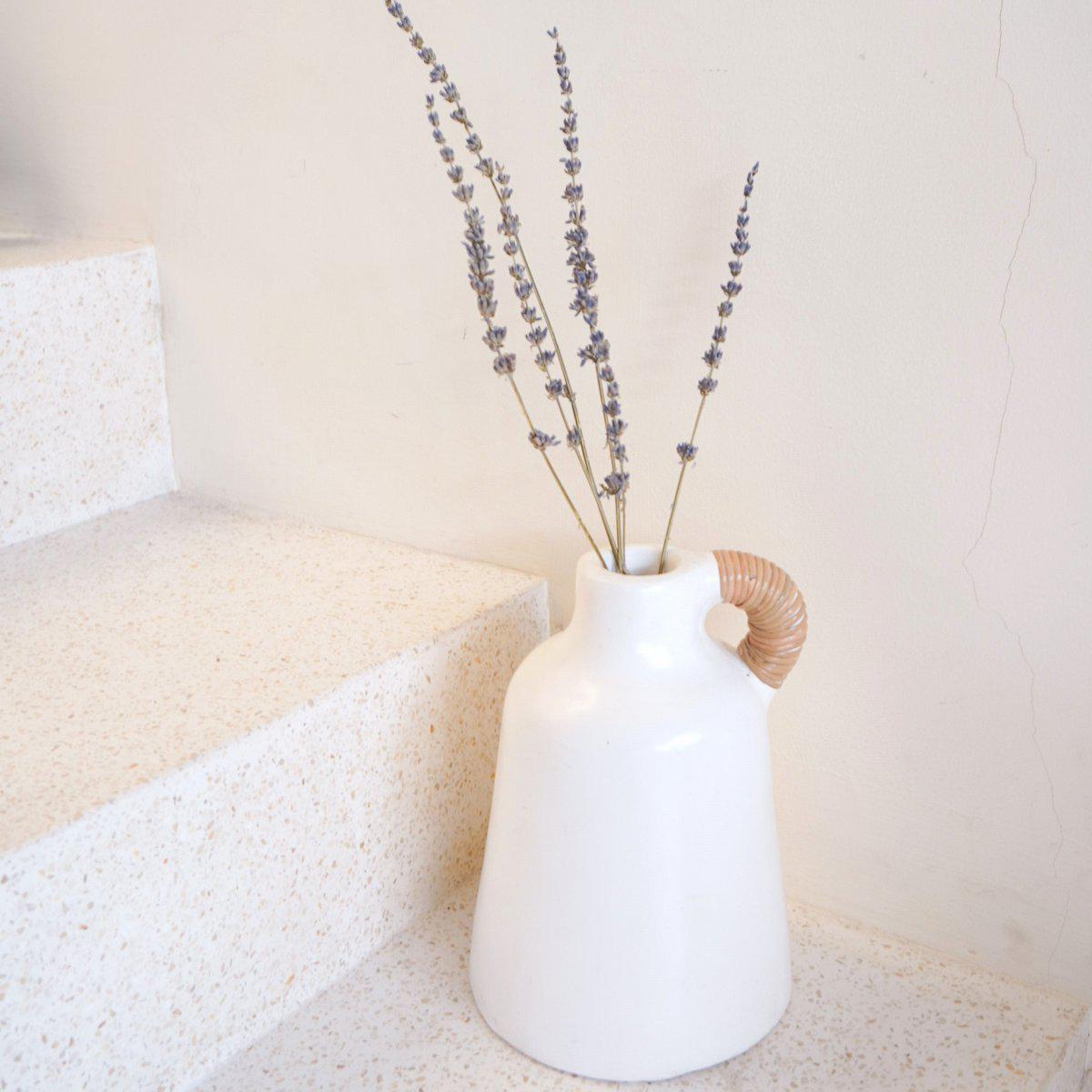 Elegante witte vaas met droogbloemen, naast een trap op een minimalistische achtergrond, met een natuurlijk touwhandvat detail.