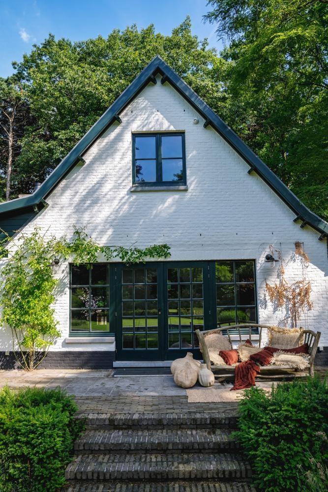 Rustieke witte cottage met groene luiken en gezellige buitenlounge te midden van weelderige bomen.