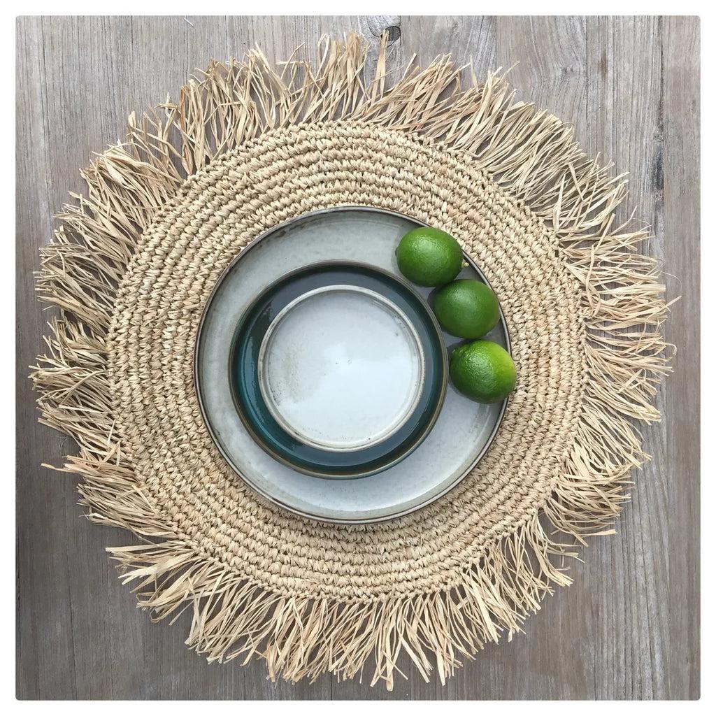 Handgeweven raffia placemat met kauri schelpen en keramisch servies op houten tafel, perfect voor een tropische tafeldecoratie.