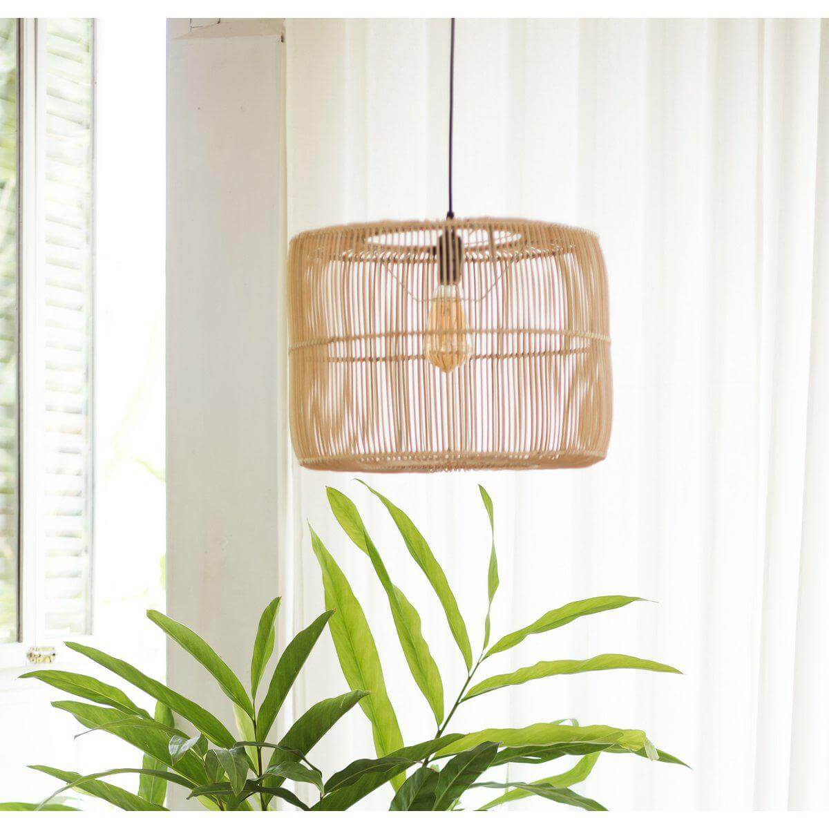 Handgemaakte rotan hanglamp LIKU in rustieke boho-stijl, met warme lichtpatronen, hangend boven groene planten in een interieur.